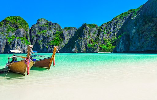 Long tailed boats are pulled up onto a golden beach lapped by green waters with a limestone rocky outcrop in the distance